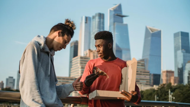 Dos personas comiendo pizza en NYC frente al skyline, simbolizando decisiones financieras comunes en EE. UU.