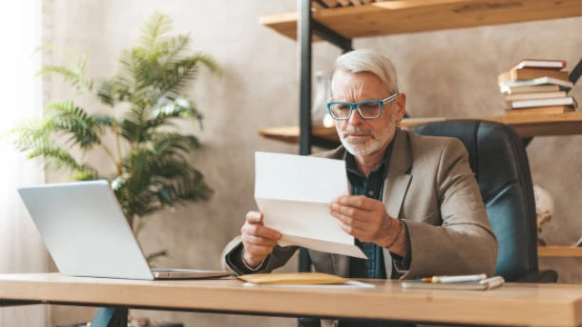 Hombre mayor en su oficina leyendo una carta con expresión de sorpresa