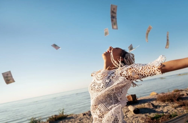 Mujer feliz y libre en la playa mientras vuelan billetes a su alrededor representando libertad financiera y tranquilidad económica.