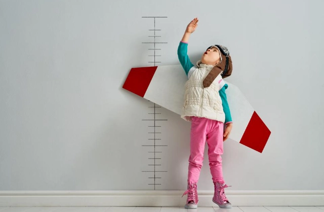 Niña midiendo su estatura con un disfraz de avión, representando altas expectativas y la posibilidad de acceder a atención médica internacional.